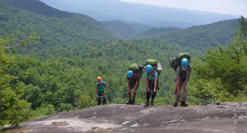 Four students wearing helmets and backpacks hold on to a rope as they scramble up a rock incline. Behind them is a mountainous landscape covered in trees.